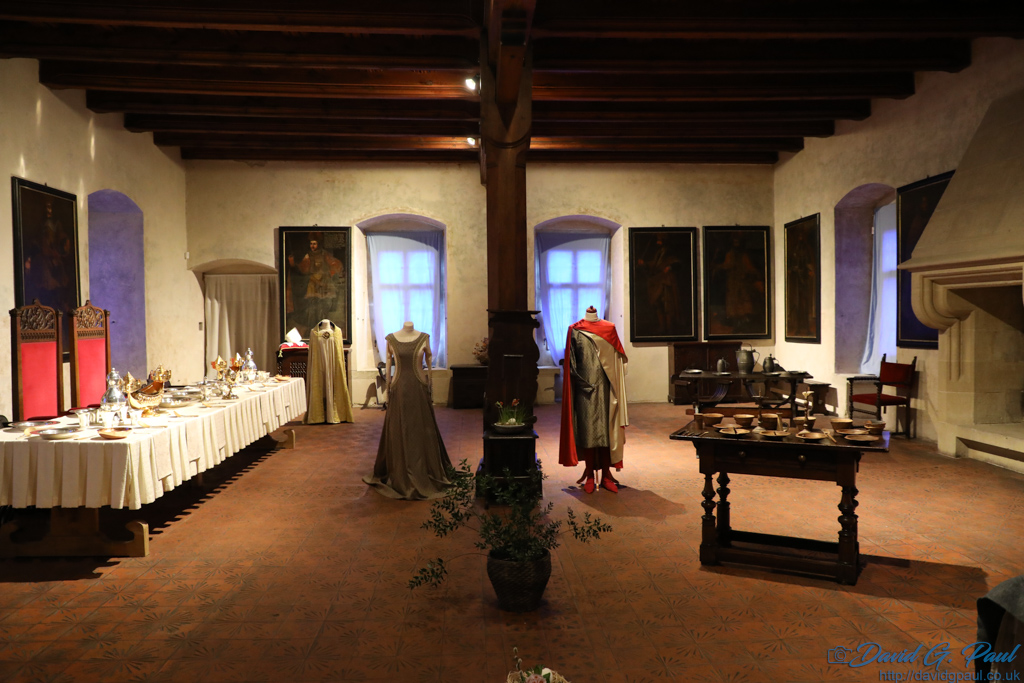 A banquet hall with two throne-like chairs, and a couple of costumes take centre. There is a large stone fireplace opposite the head table.