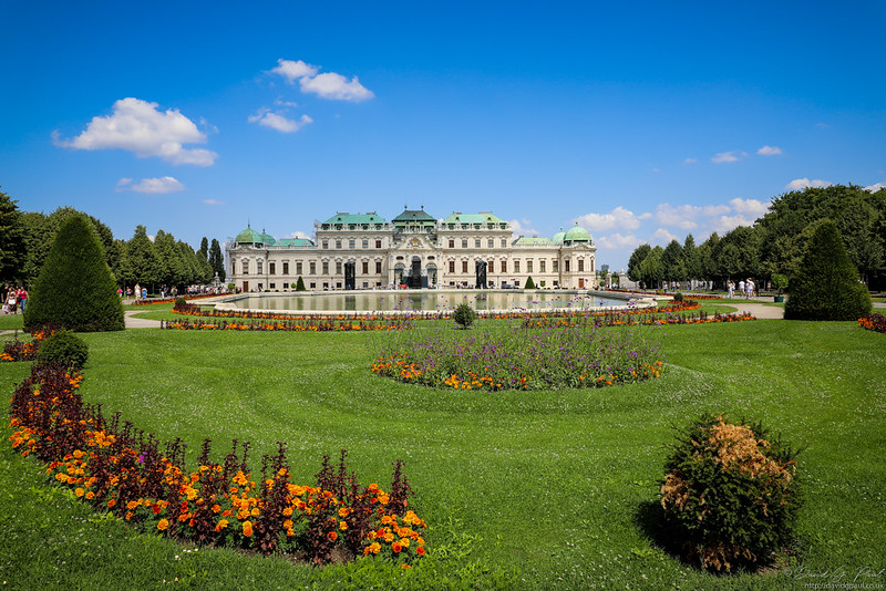 A large white building with a light green roof sits behind a large body of water surrounded by carefully shaped trees. In front of it are colourful flowers.