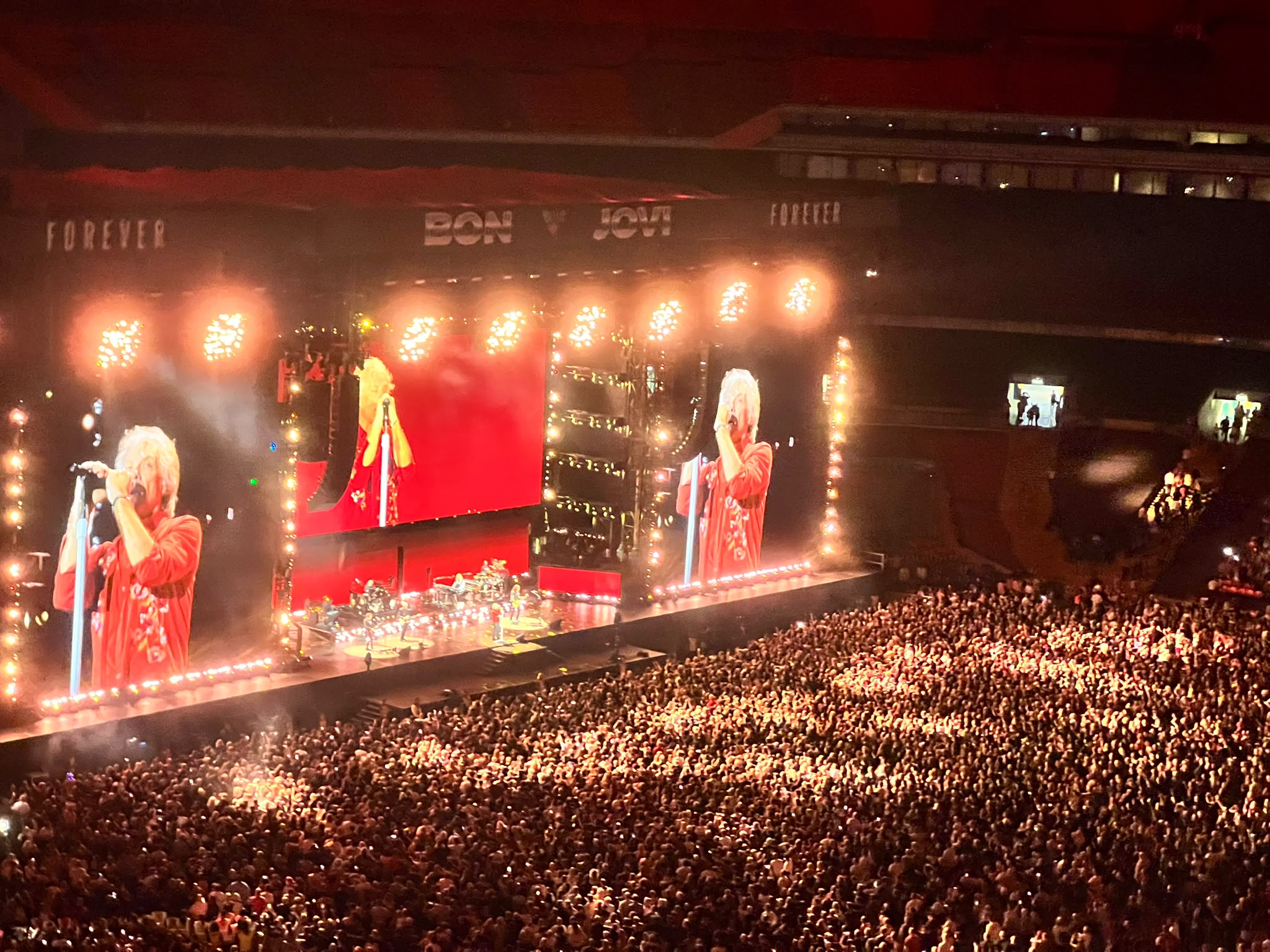 Massive stadium crowd watching Bon Jovi perform live on a brightly lit stage with giant LED screens.