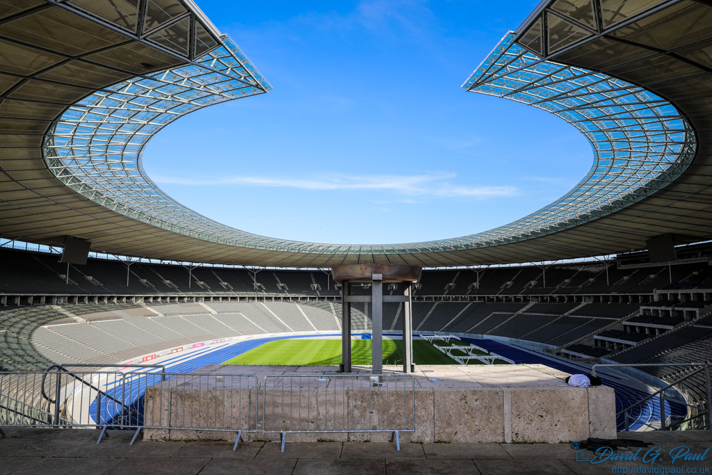 Olympic Flame Bowl, and beyond you can see seating, a blue running track around afield, and above is a roof with a cutout above the bowl